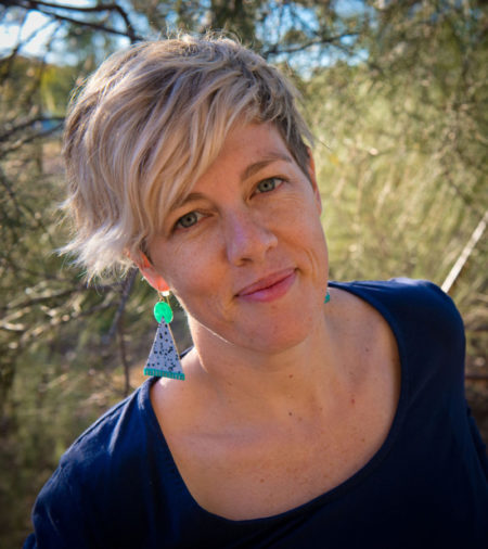 Alex Kelly Headshot Alex in a black top with short blonde hair with bright earrings by_Anna Cadden