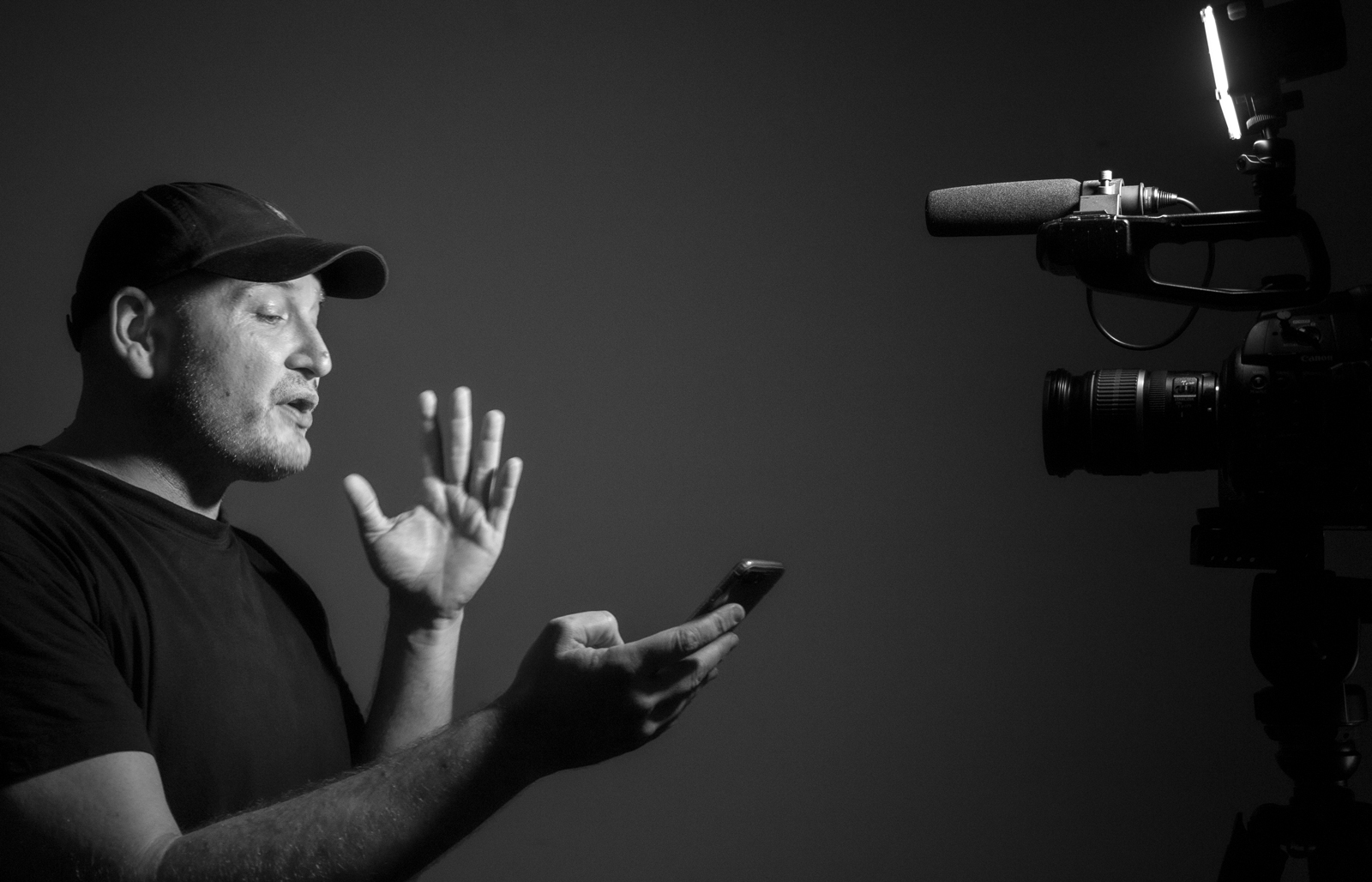 Emile Zile Headshot Emile in front of a camera holding a phone in black and white talking by J. Hall