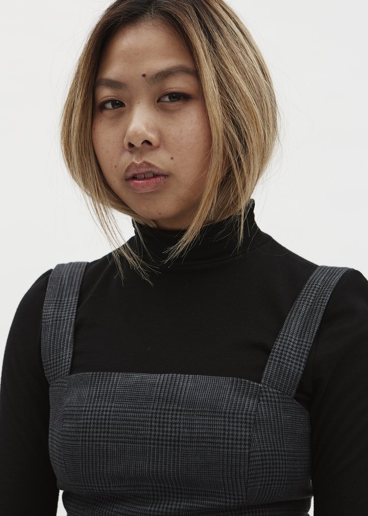 A portrait of a Chinese-Australian woman in her mid 20's with mousy blonde hair. She is wearing a black long-sleeved turtleneck and chequered grey and black overalls.jpeg