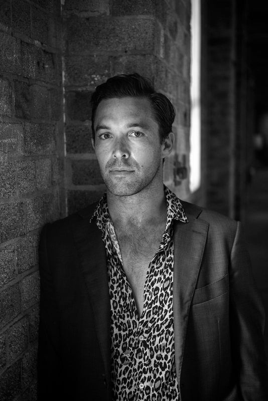 Black & White Image of a man leaning on a brick wall wearing a leopard print button up shirt layered with an unbuttoned suit jacket on top.
