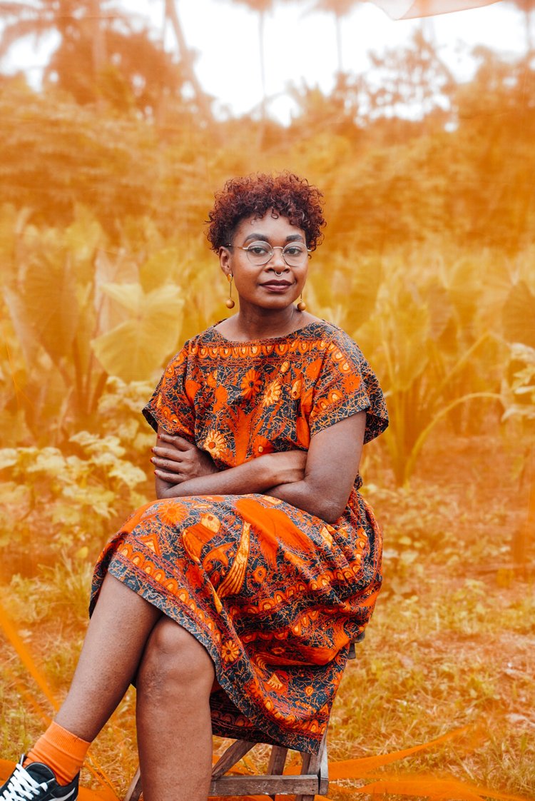 A portrait of a Tokelaun-Fijian woman in her late 20's with loose curly short red hair with a side cut. She is sitting upright on a chair in front of a see-through tinted orange backdrop. She is wearing eye glasses, orange earrings and a patterned orange and black dress