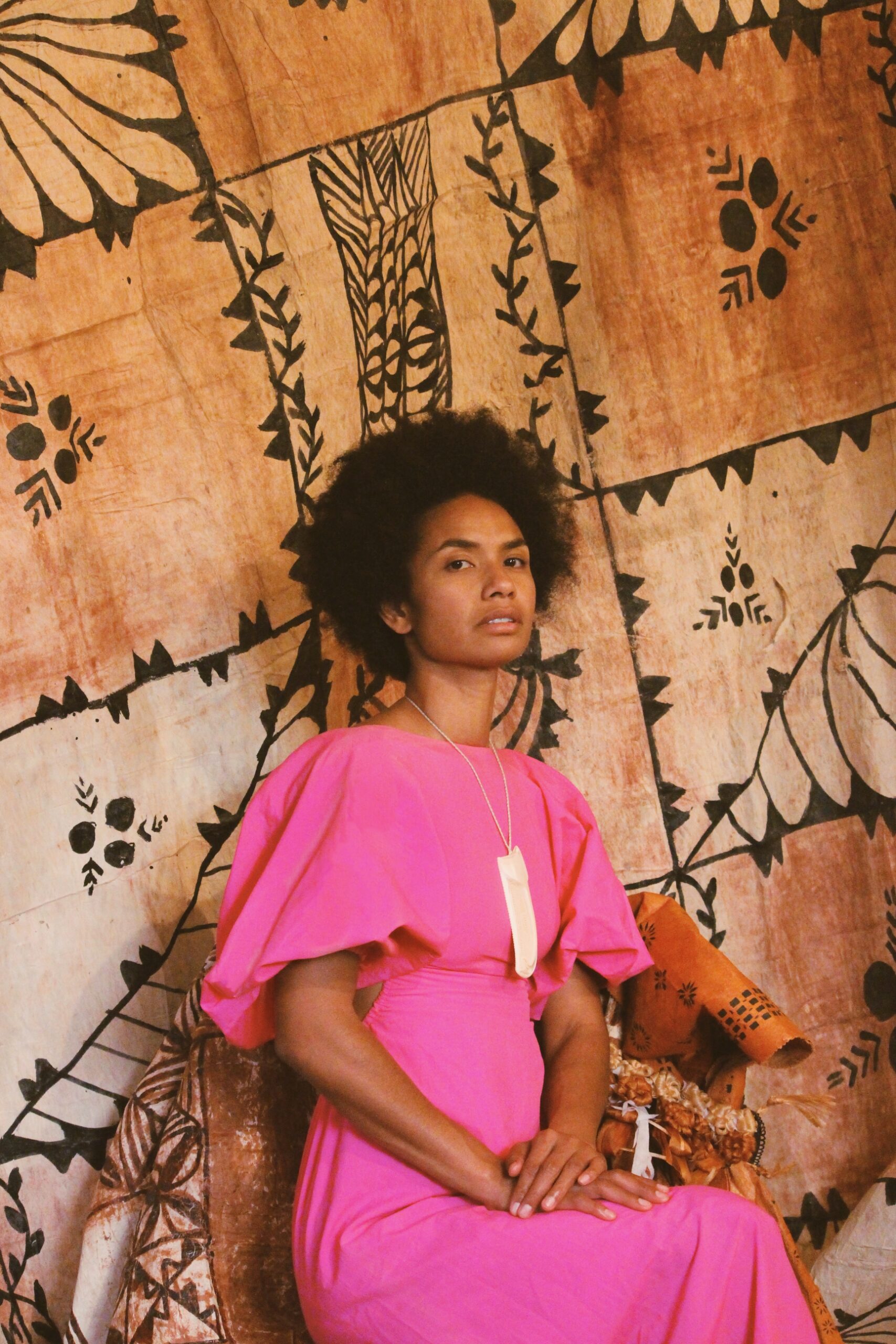 A portrait of an Itaukei woman in her late 20's with a jet black buiniga hairstyle. She is sitting upright on a chair in front of ngatu. She is wearing a pink dress with a necklace
