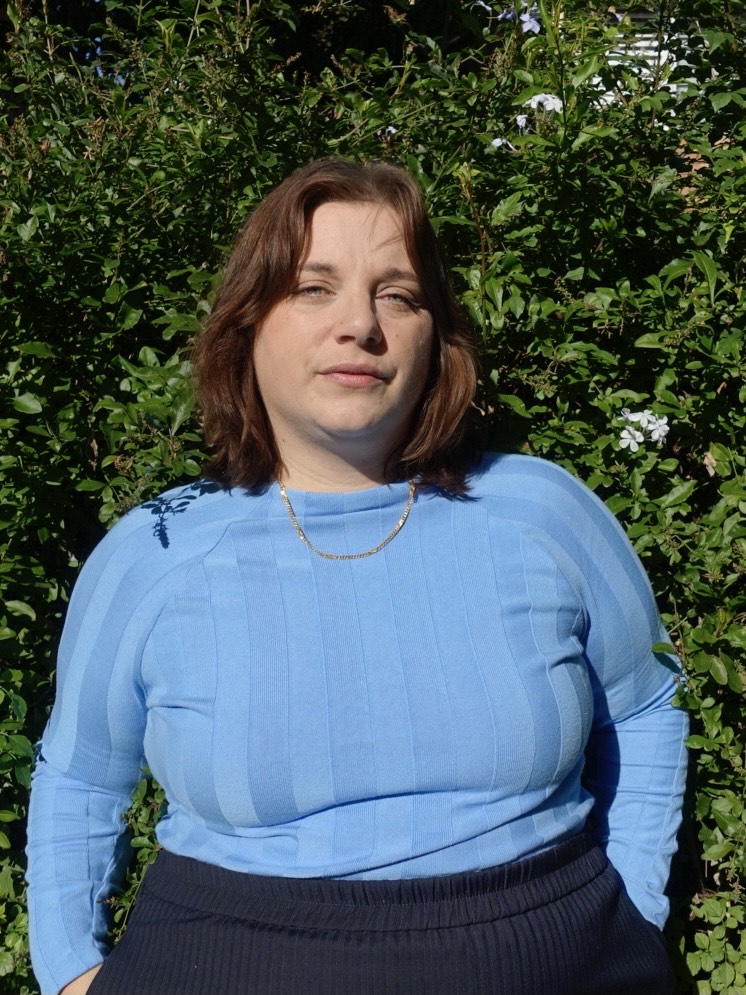 Image of a woman in a blue shirt, with fair skin, dark hair and blue eyes with a background of green shurbbery.