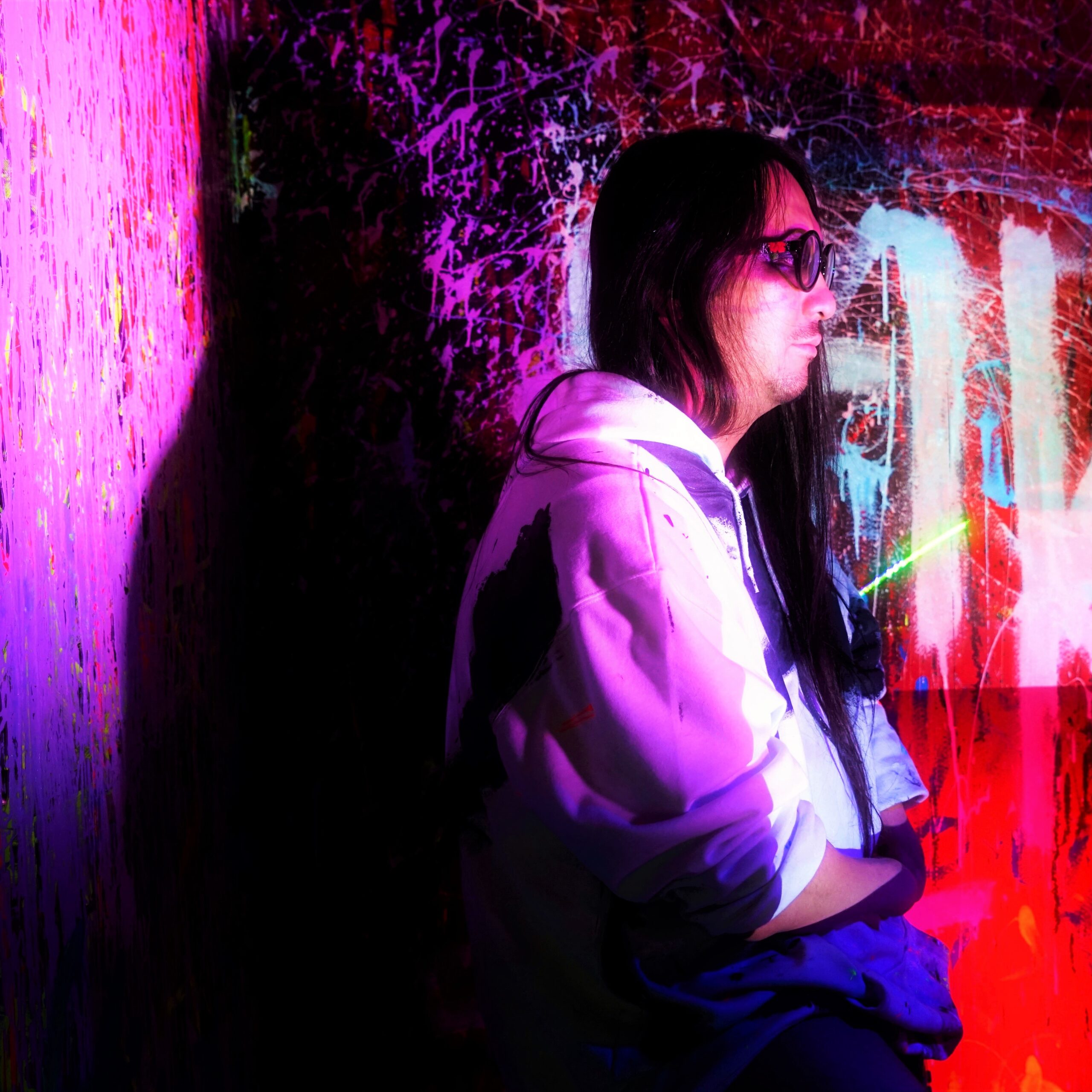 A colourful image of a person with long black hair and glasses facing sideways infront of graffiti coloured walls