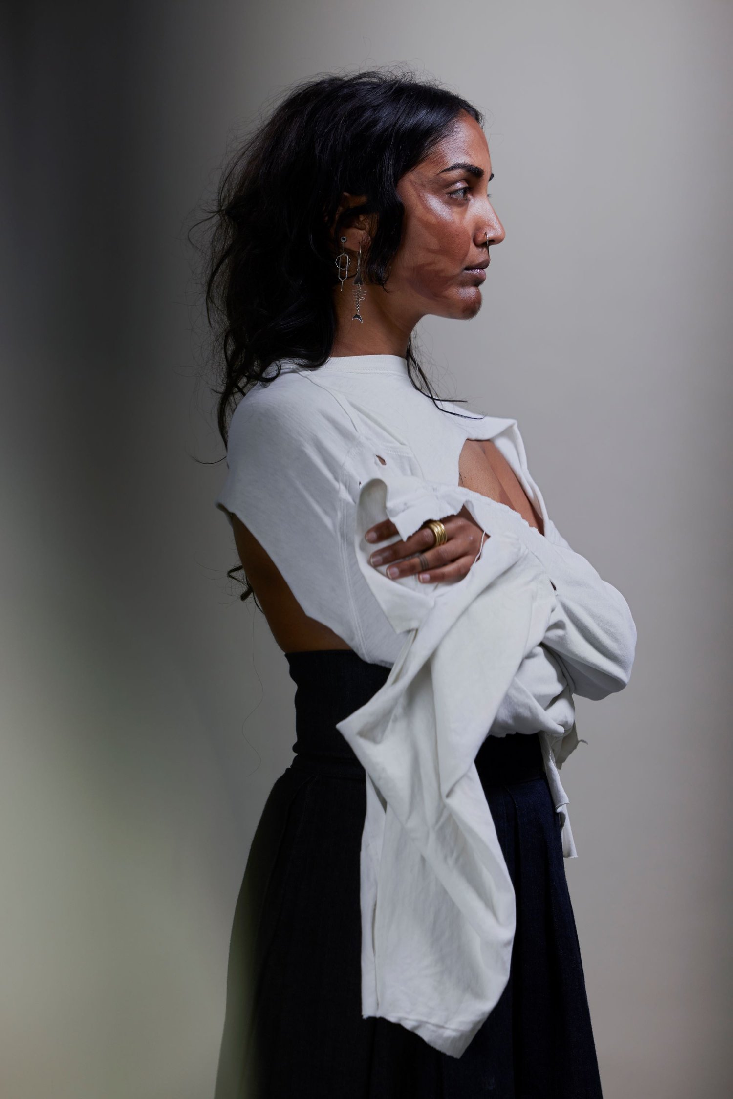 Person with shoulder-length hair wearing a billowing backless white blouse stands with their arms crossed in front of a shadowy wall. They are facing the side, revealing the henna on their face and dangling earrings.