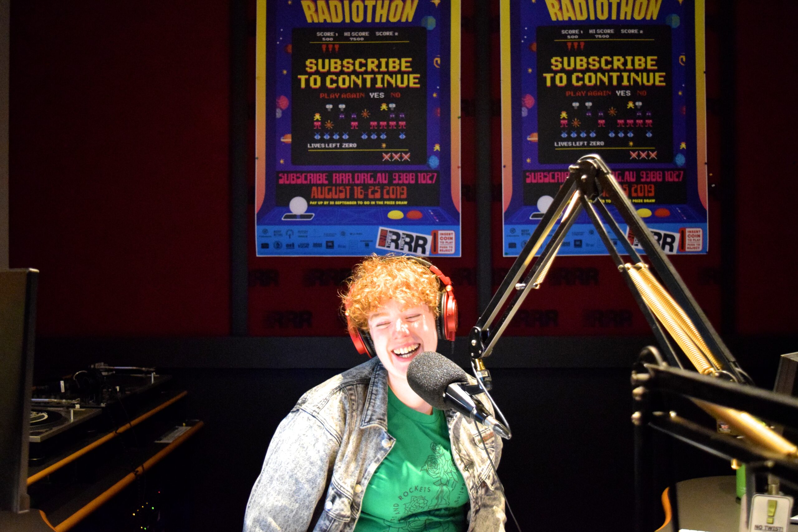 Person with short curly red hair and green shirt laughing in front of a mic at the Triple R radio station studio.