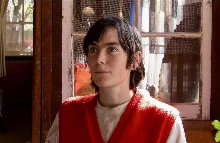 Person with short brown hair wearing a red sweater vest gazes off camera as they sit in front of a rustic window and hanging fabrics.