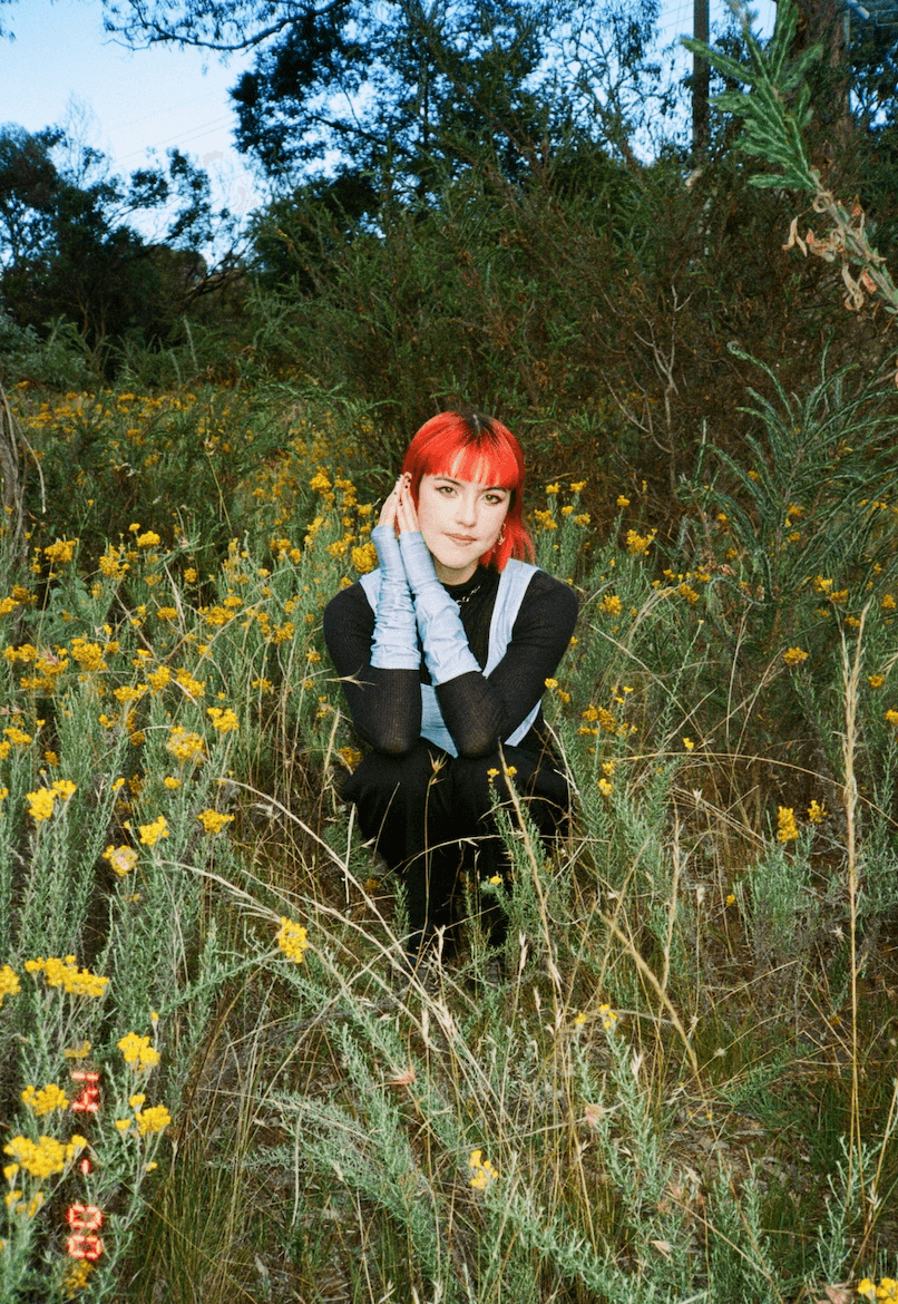 Person with bright red-orange fringe squatting in a field of weeds sprouting yellow flowers. They wear a long-sleeved black outfit accented with light blue vest and matching fingerless gloves.