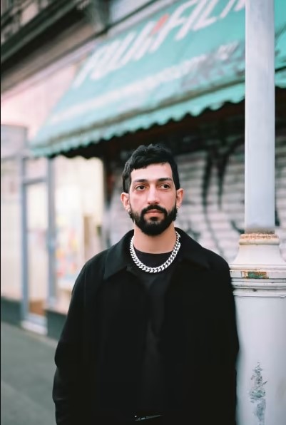 Person with short black hair and thick beard wears a black jacket and silver chain as they lean against a street pole in front of blurred storefronts.