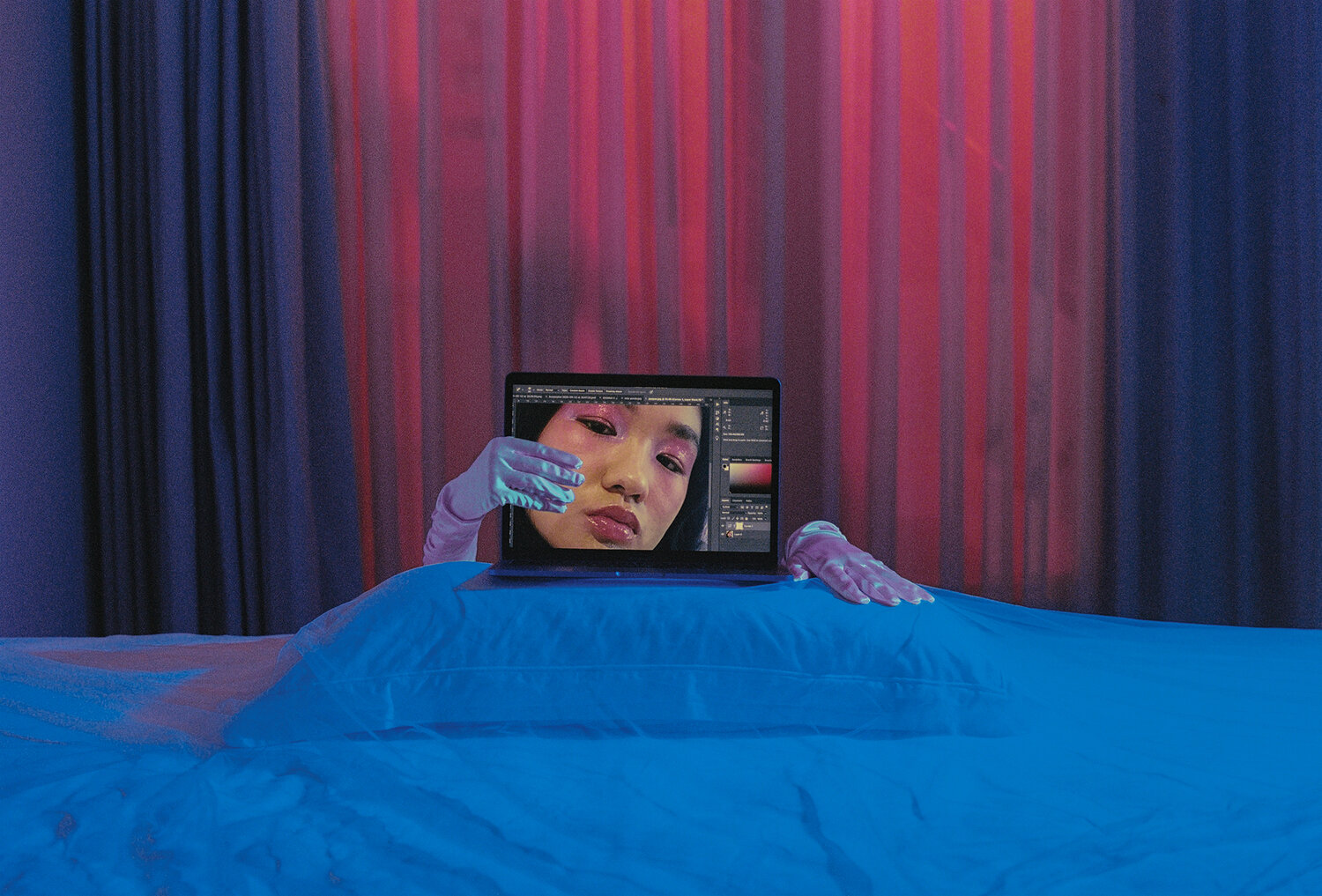 Laptop screen opened to a zoomed-in image of a sultry face with black hair, pink eyeshadow and lip gloss. The laptop is in a neon blue and pink bedroom, its screen held by hands in white satin gloves.