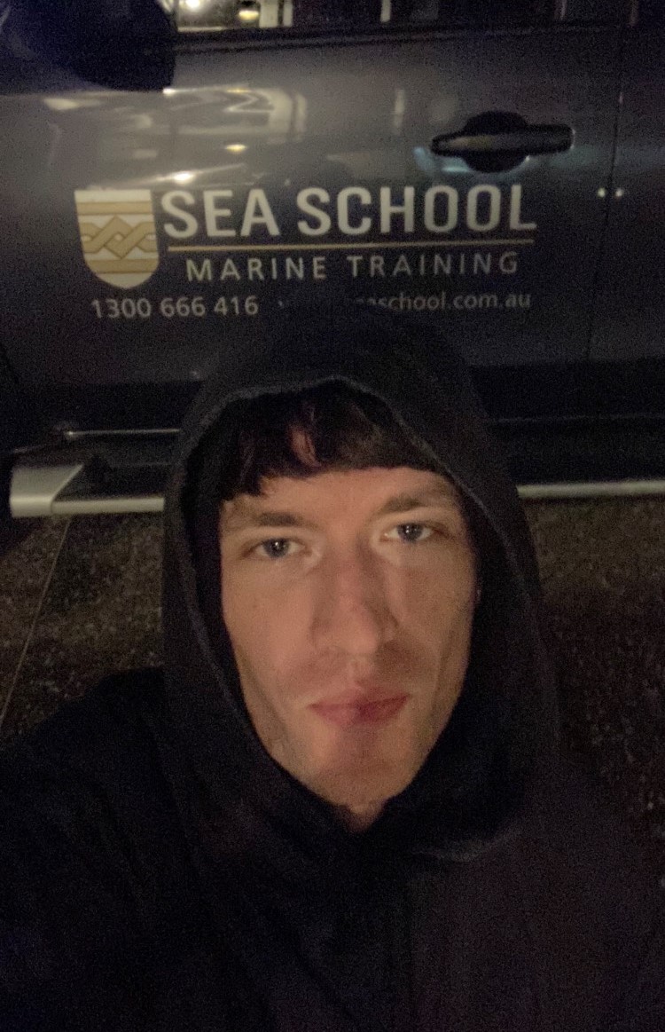 Selfie of person with short dark fringe and black hoodie in front of a parked car advertising “Sea School marine training”.