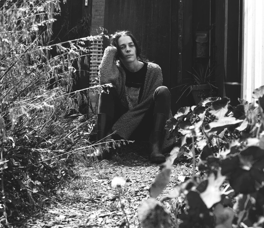 A black and white image of a person sitting in a backyard, surrounded by foliage. They have medium length dark hair and wear an oversized cardigan with gumboots.
