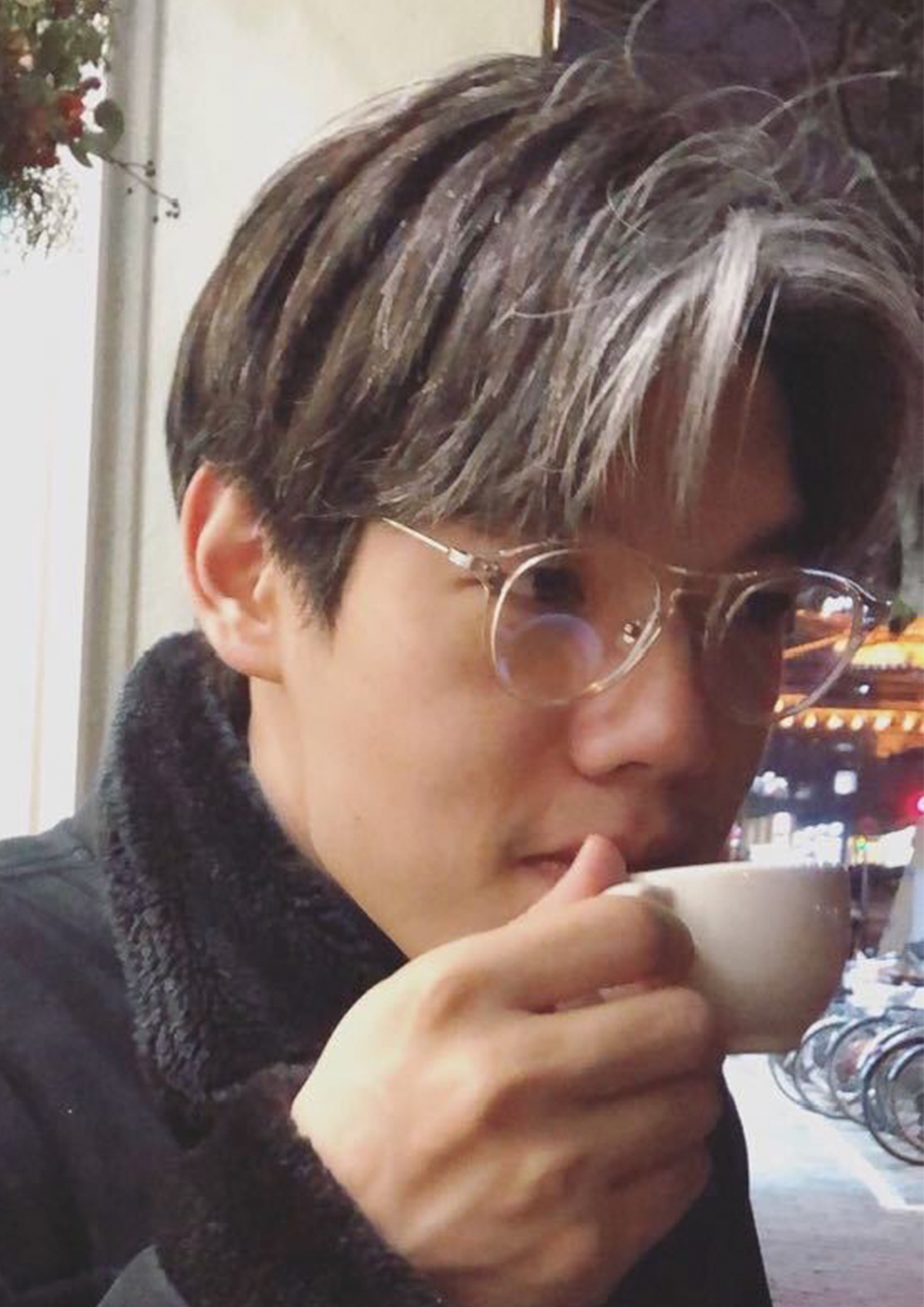 This image shows you a young artist with marron and white colour hair, wearing a pair of glasses. He was wearing a black winter coat with fur collar , drinking espresso.