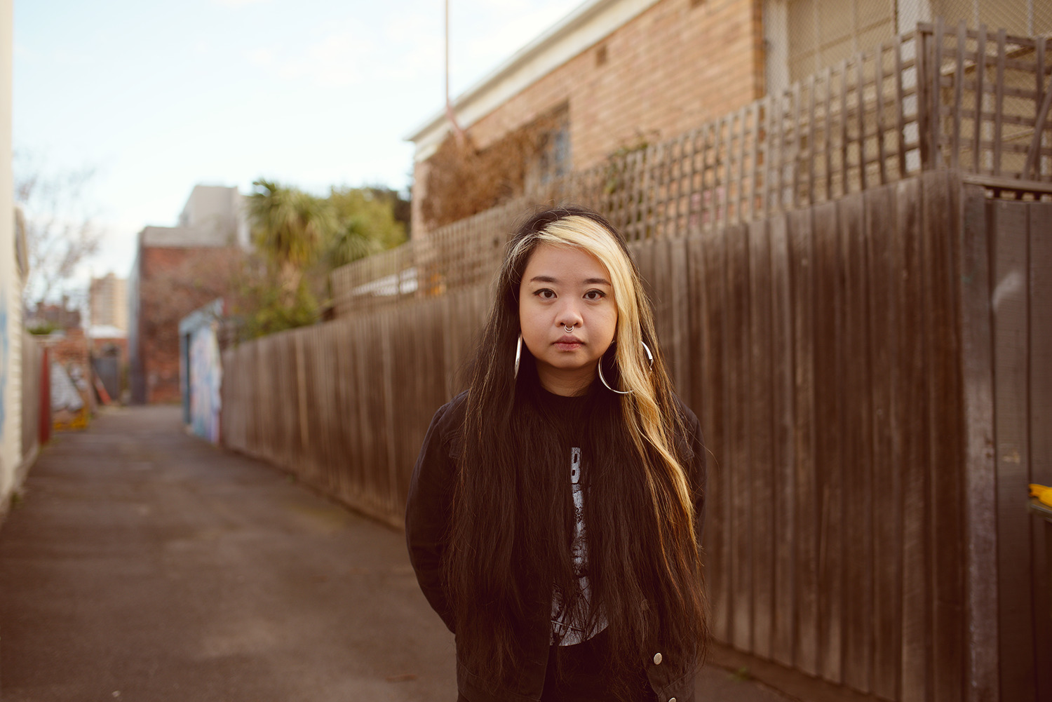 A woman stands in a laneway with long black hair and a streak of blonde down one side looking directly at the camera