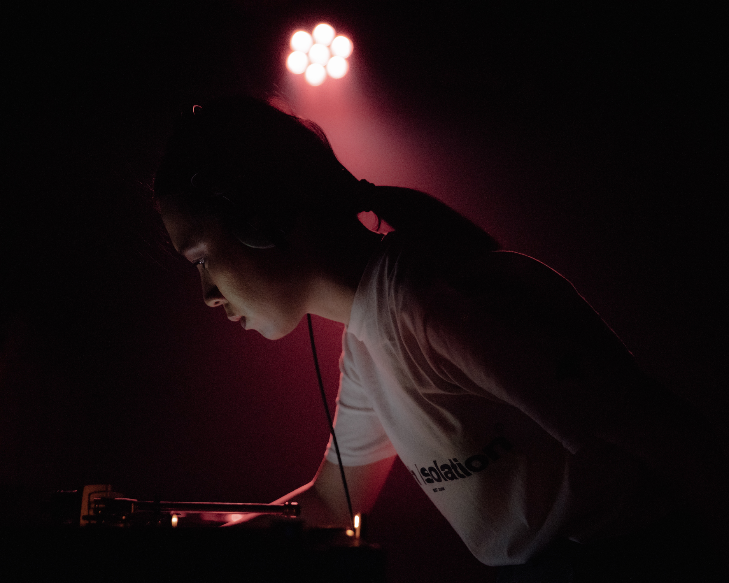 Portrait of a woman looking away, at a DJ table performing a set. She is wearing a white tshirt and has her hair in a ponytail