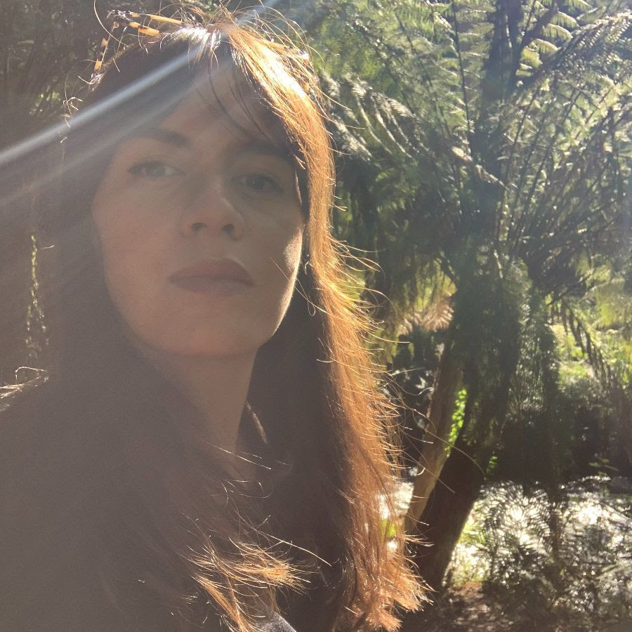 A woman with long dark hair is looking at the camera. There are lush plants behind her and sunlight streaming across her face.
