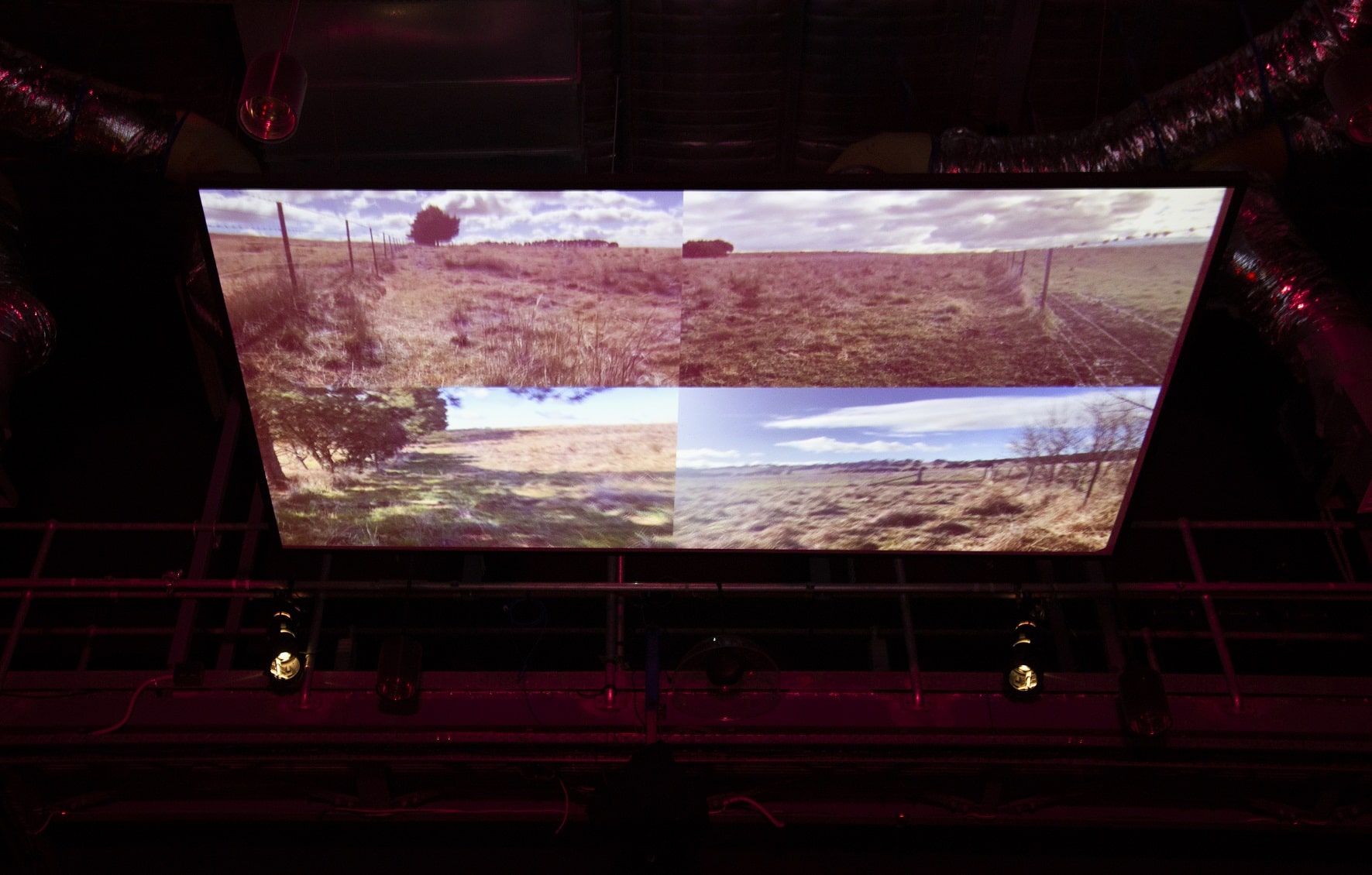 A large projection screen is suspended in the industrial ceiling surrounded by pipes and technical equipment. Projected on the screen is four seperate images in each quater of the screen, each displaying imagery of a rural country setting with brown unmaintained grass and weeds, blue sky boparded by barbed wire fences.