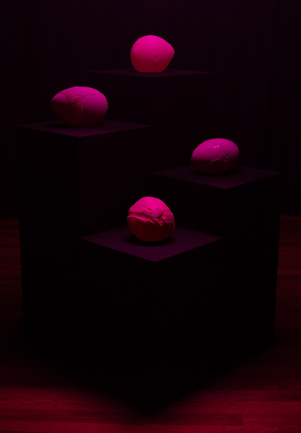 A darkly lit room, with woodeen floor boards and black curtains with four black plinths of different heights centered in the middle with four different bright pink blobs centered on each plinth. Each of the blobs have distored images of faces and Chinese karaoke characters.