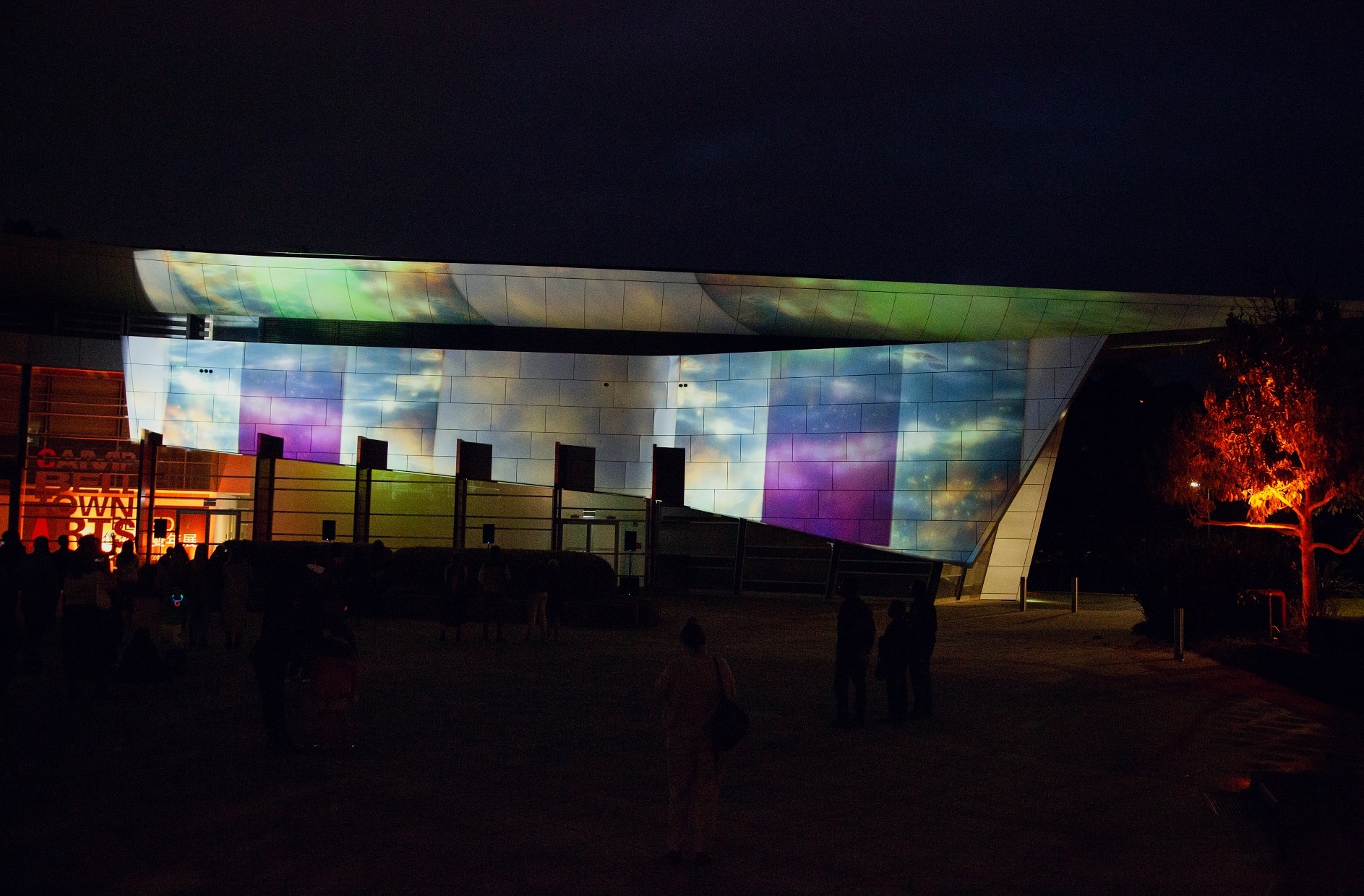 The Campbelltown Arts Centre building with sharp archictectural lines and a rectuacular screen like panel has imagery of glowing purple pillars with a background of light reflecting on light blue rippiling water and a light blue sky is projected onto it, with the dark night sky the backdrop to the building.