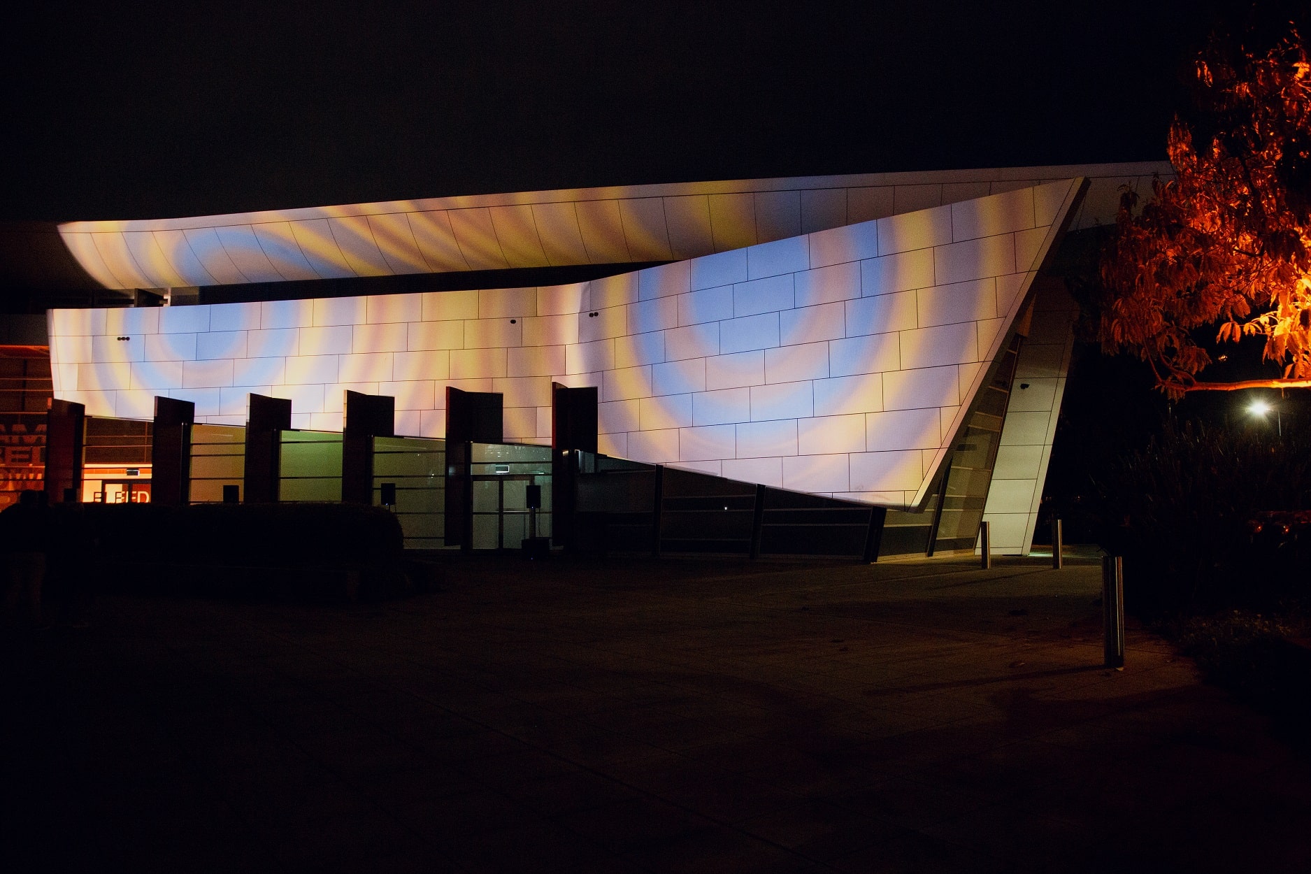The Campbelltown Arts Centre building with sharp archictectural lines and a rectuacular screen like panel has rings of deep light blue, grey and orange rings projected onto it, with the dark night sky the backdrop to the building.
