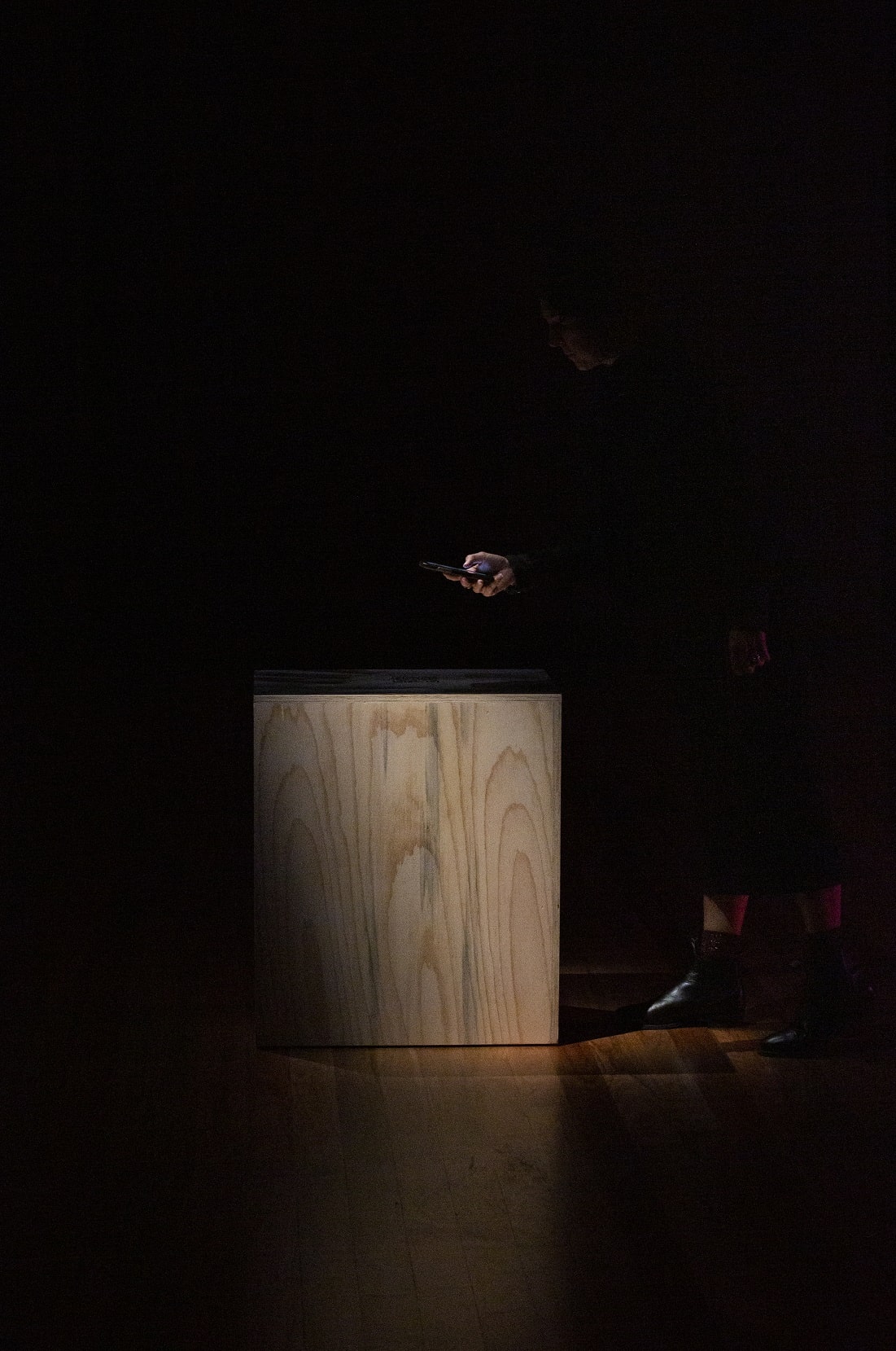 Dark image or a wooden plinth from the side and a person standing, obsured by shadows. They stand over the plinth with their hands and phone looming out of the dark shadows to capture the QR code image on top of the plinth.