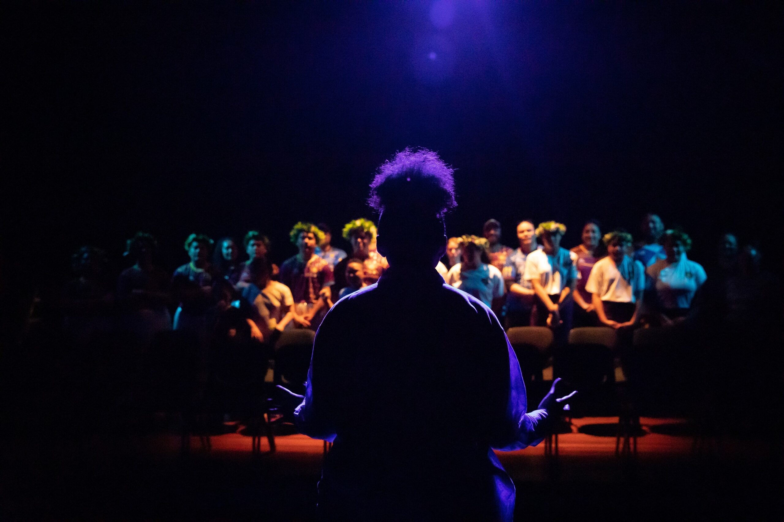 A person with their back to the camera stands infront of a seating bank full of young members of a choir. The lighting is blue.