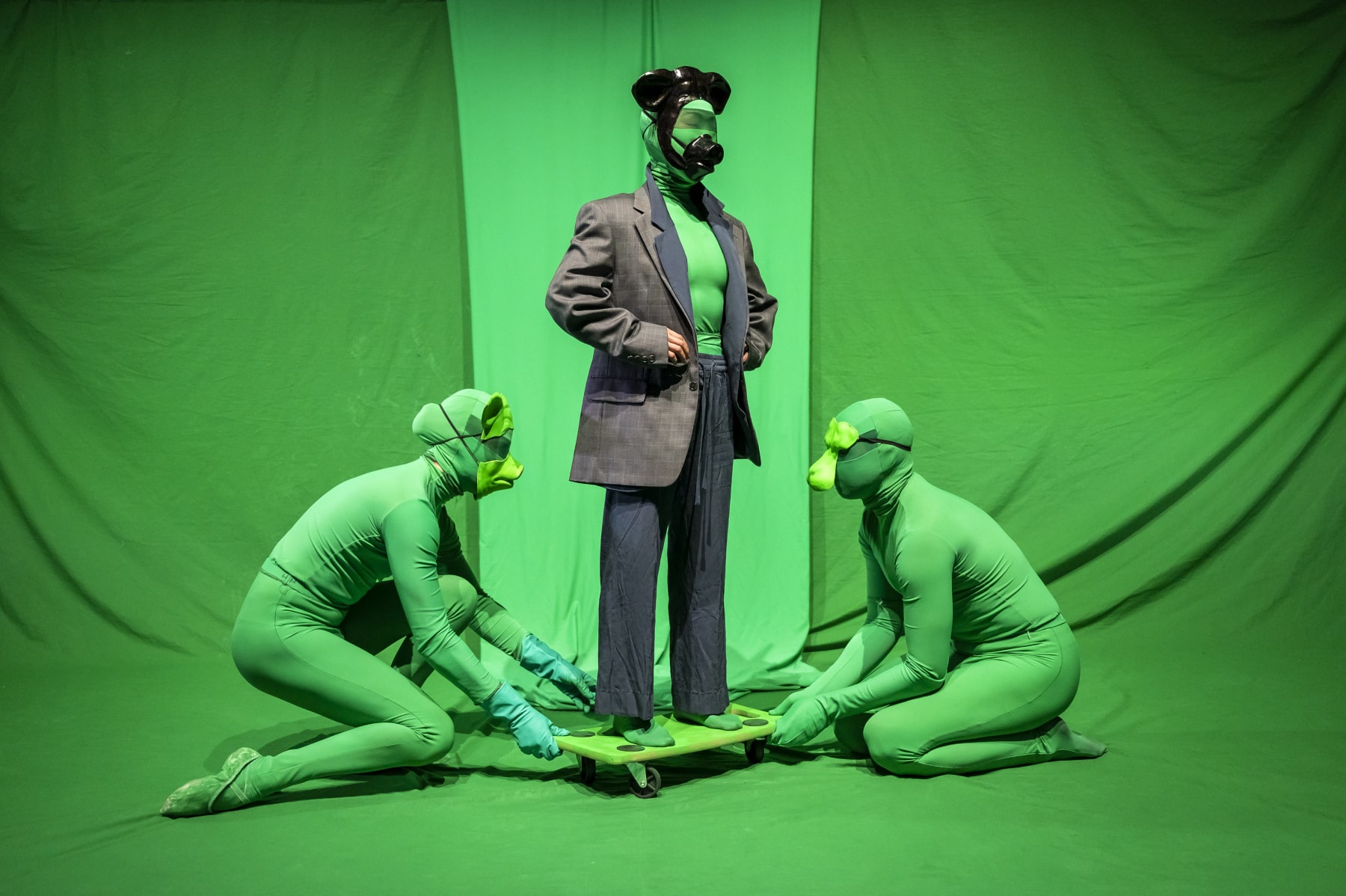 Three people are in front of a green screen dressed in skin-tight green suits and animal masks. The person in the middle wears a grey suit over the top and is standing on a green trolley. The two other people are crouched on either side of them holding the trolley.