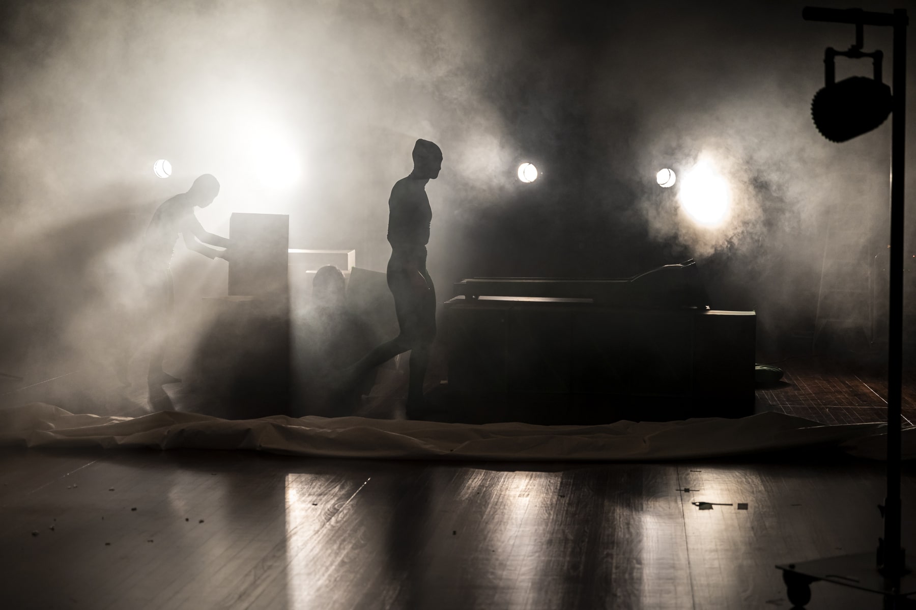A stage with bright white lights and haze. You can see two figures in the haze. One is lifting up a box, the other is standing beside a tredmill.