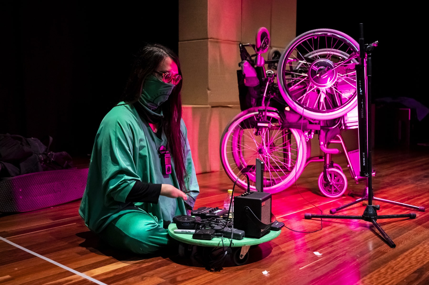 A person is sitting on the floor wearing green. They have long dark hair and glasses. In front of them is music mixing equipment. In the background there is a wheelchair that is lit up with pink lights and a stack of cardboad boxes.