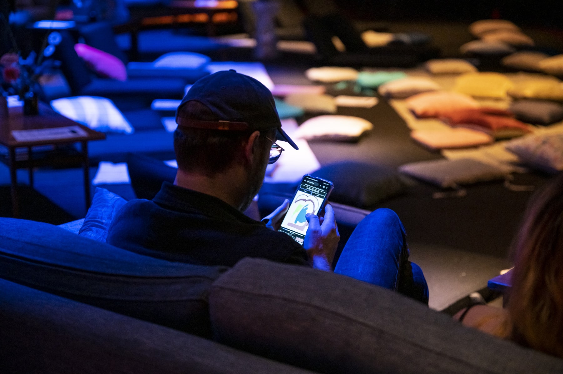 A person wearing a cap and glasses is drawing on their mobile phone. They are sitting on a couch and their back is to the camera. In the background there are cushions on the floor. There are coloured lights.