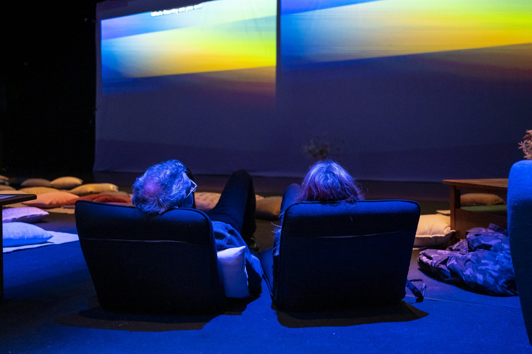 Two people are lying on black lounge chairs. Their backs are to the camera. They are watching two large screens with an gradient of colours on them.