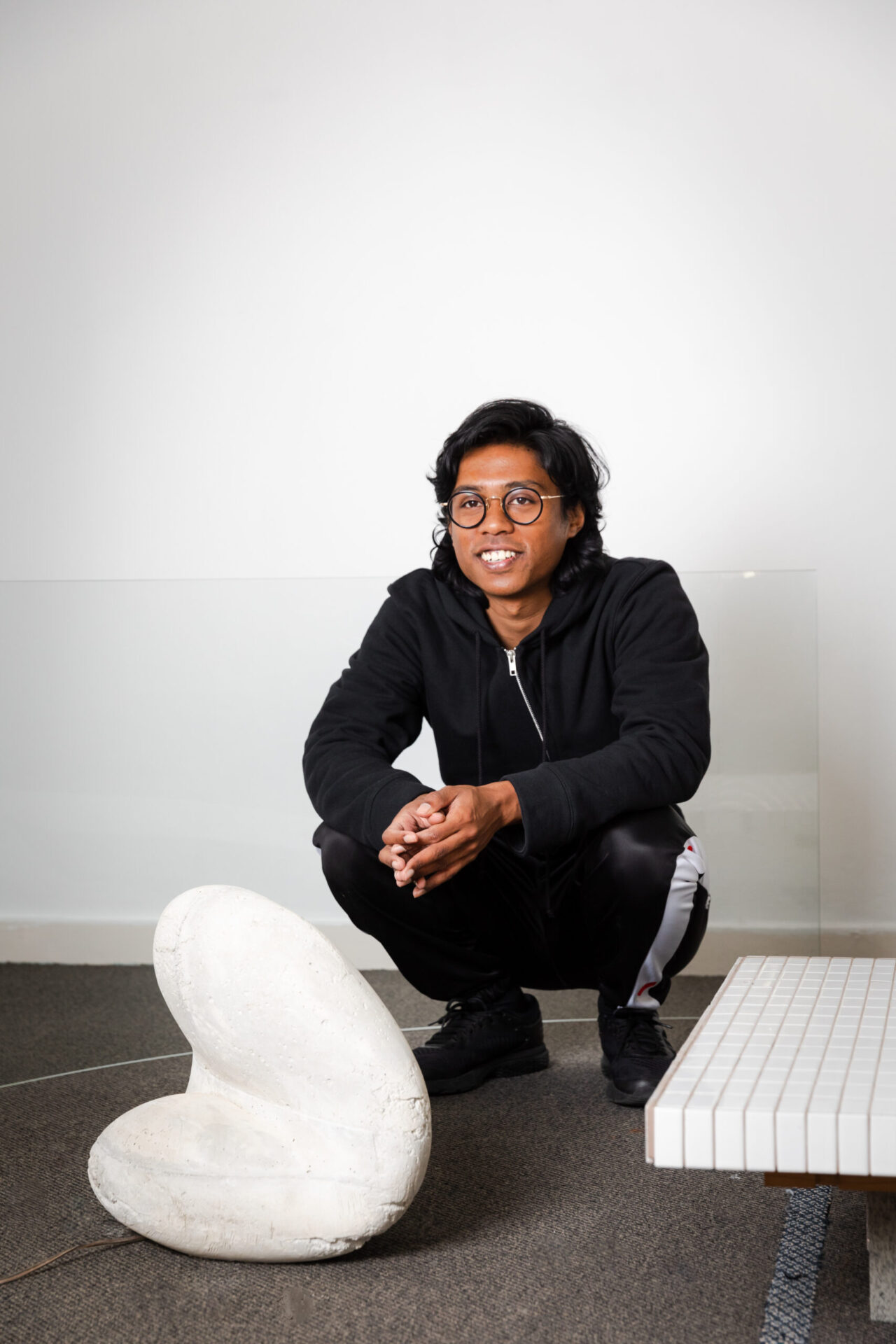 Akil is crouching on the floor indoors, positioned behind a solid curved white sculpture and wearing a black tracksuit and round framed glasses.