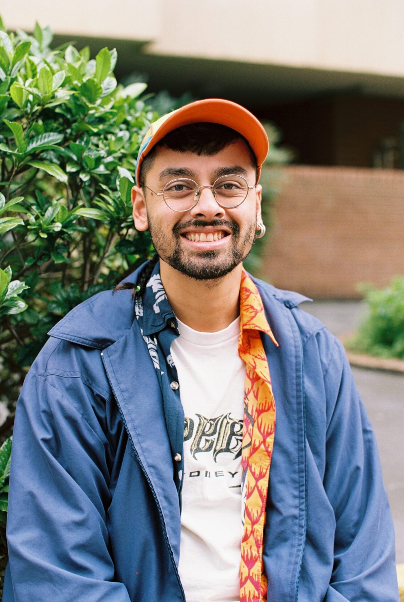 Kalanjay is smiling at the camera, standing outdoors in front of a leafy bush and an out of focus brick wall. He wears round framed glasses, an orange hat and layered shirts of white, orange and blue.