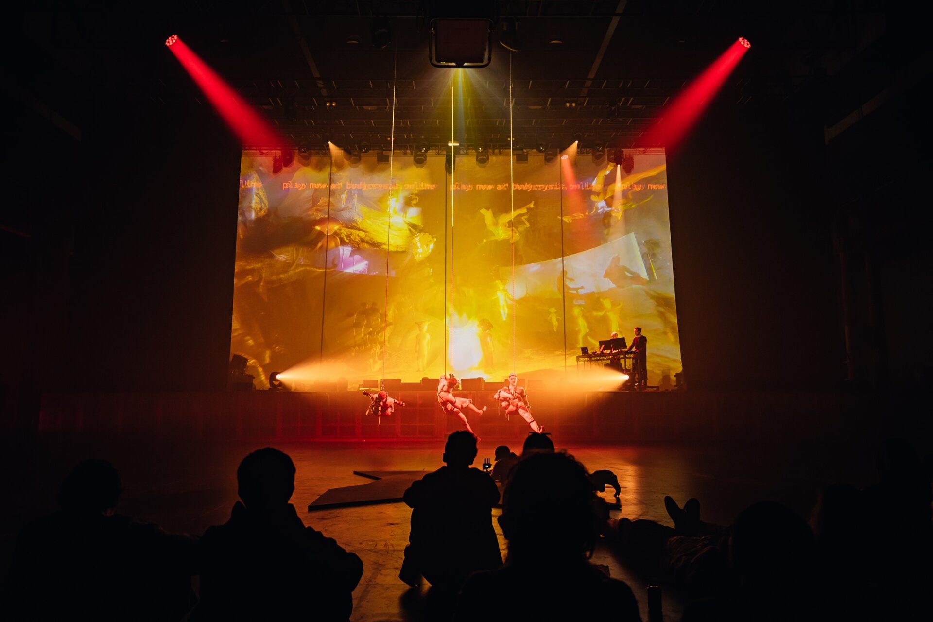 A crowd views three dancers suspended from the ceiling performing movement in front of a large screen filled with 3D animated avatars.