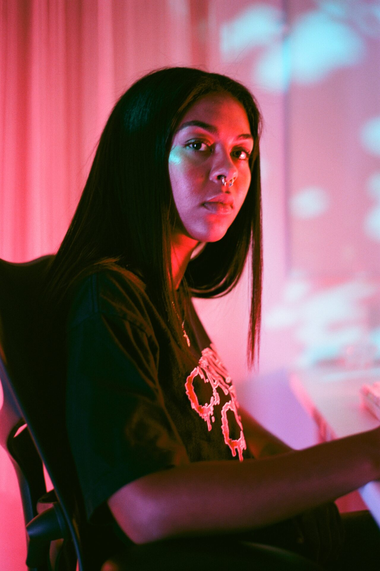 Serwah turns to face the camera as she sits on a black computer chair at a white desk (out of shot) in a soft pink room with a vibrant pink, red and turquoise light reflecting on her face. Serwah has long dark straight hair and wears a gold chain around her neck, a septum piercing and a black t-shirt with dripping red text on the front.