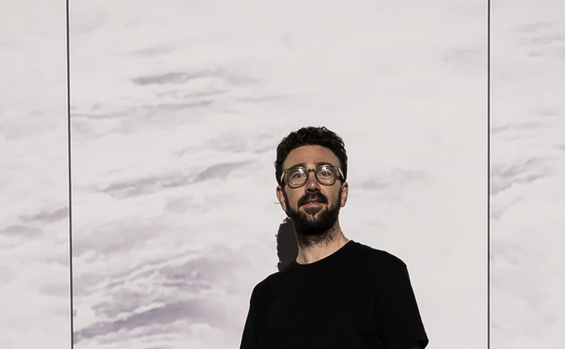 An image of artist Tom Blake from the mid-chest upwards. He is white, with dark brown hair, a beard, moustache, and black glasses. He is staring directly at the camera and smiling slightly. He is standing in front of a projected image of a light background, so his face is illuminated and you can see his dark shadow behind him.