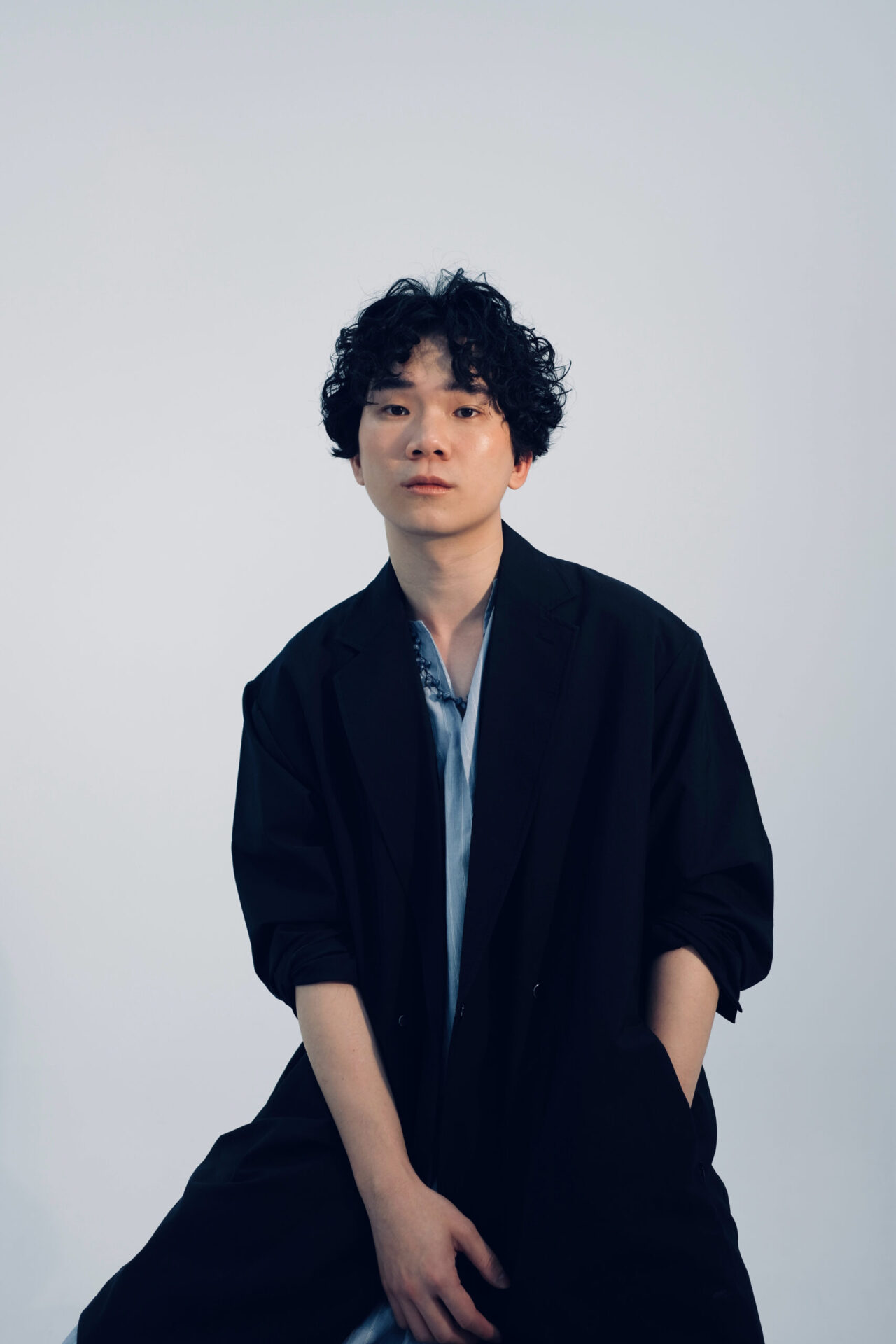 Artist Yinga Chen is seated on a stool and his body is visible from the knees up. He is wearing black pants with a black jacket and a blue shirt underneath. His hand is in his pocket. He is looking directly at the camera with a serious expression on his face. His hair is black and curly.