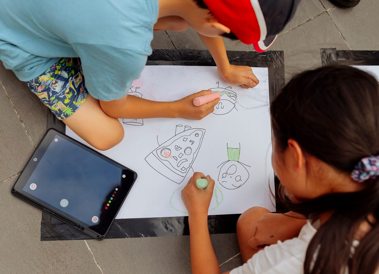 Image shows two children kneeling on the ground drawing on a large piece of paper using coloured chalk with an iPad placed beside them on the left. The children are leaning over the page as they are drawing. One child has a red and black hat, blue short sleeve t-shirt and patterned shorts. The other child is wearing a white t-shirt and has long dark straight hair tied up in a ponytail with a purple, blue and pink patterned scrunchie.