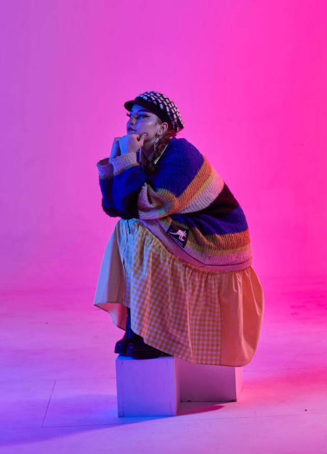 Anouk is sitting on a plinth with her feet resting on a smaller plinth facing the left, with her arms bent and resting on her knees and hands resting under her chin. She is sitting in front of a pink tinted background. She is wearing a black and white beanie which has a small cap at the front, a jumper with blue, orange, yellow, white and black stripes and a yellow checked dress underneath.