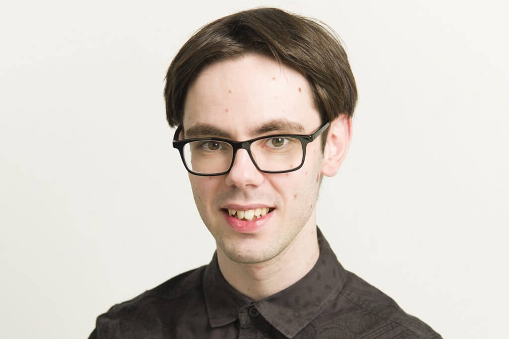 Blake is smiling at the camera, sitting in front of a light grey background. He wears dark framed glasses, has short dark brown hair and is wearing a dark brown paisley patterned shirt.