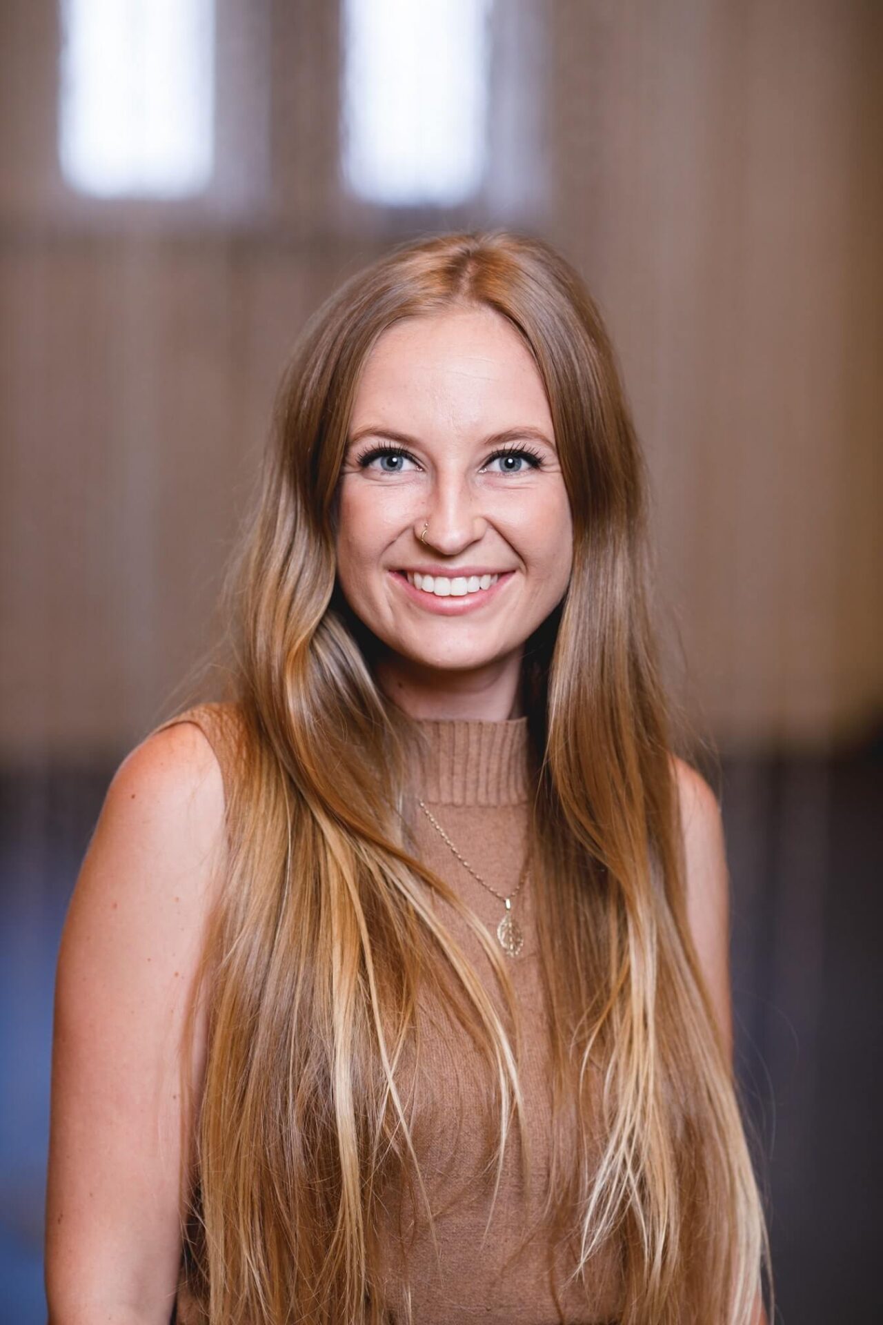 A woman with long blonde hair smiling with a blurred background of a room with an indistinguishable hanging artwork behind.