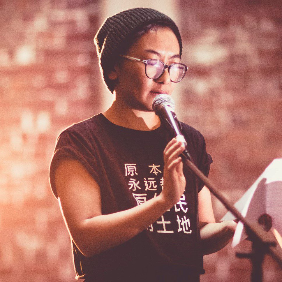 A photo of Jinghua Qian performing spoken word, wearing a beanie, glasses and a t-shirt that says 