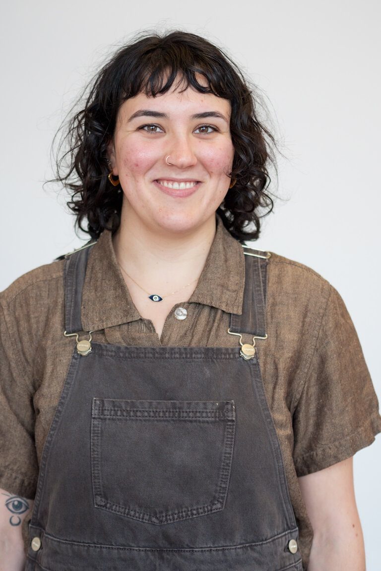 Laila is smiling at the camera, and is in the middle of the frame white a white background behind her. She wears a brown relaxed collared shirt with black overall over the top, and she has fair skin and short dark curly hair.