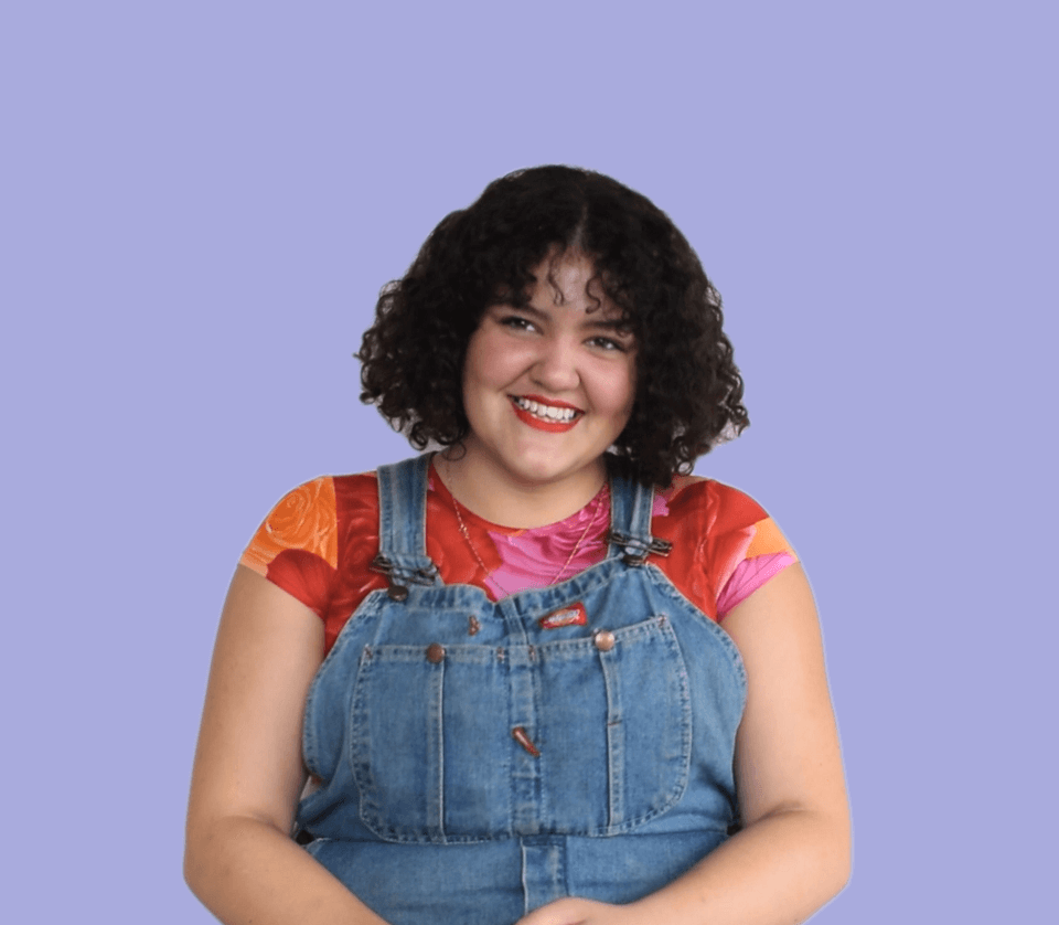 Rosemary is smiling at the camera, sitting in front of a light purple background. She has mid-length dark curly hair with a fringe. She is wearing a short sleeve floral t-shirt in red, pink and orange and denim overalls over the top.