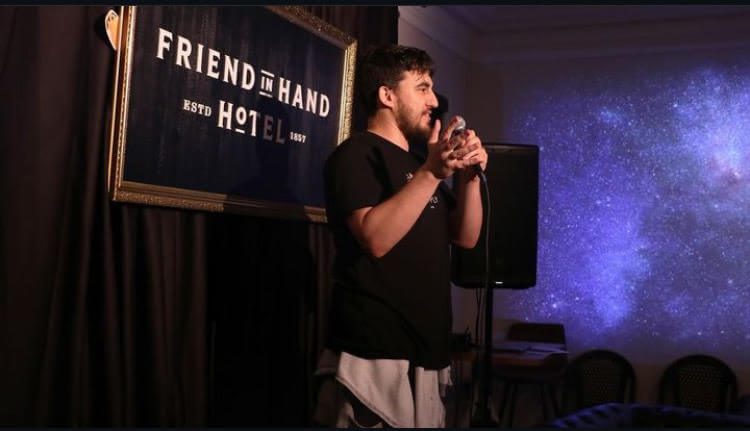 Sam is standing side on, facing towards the right. He is talking, holding a microphone in one hand and his other hand is open, directed towards the left. He is standing on stage in front of a black curtain and speakers. He has short dark hair and a neat beard. He is wearing a black short sleeve t-shirt and light coloured pants.