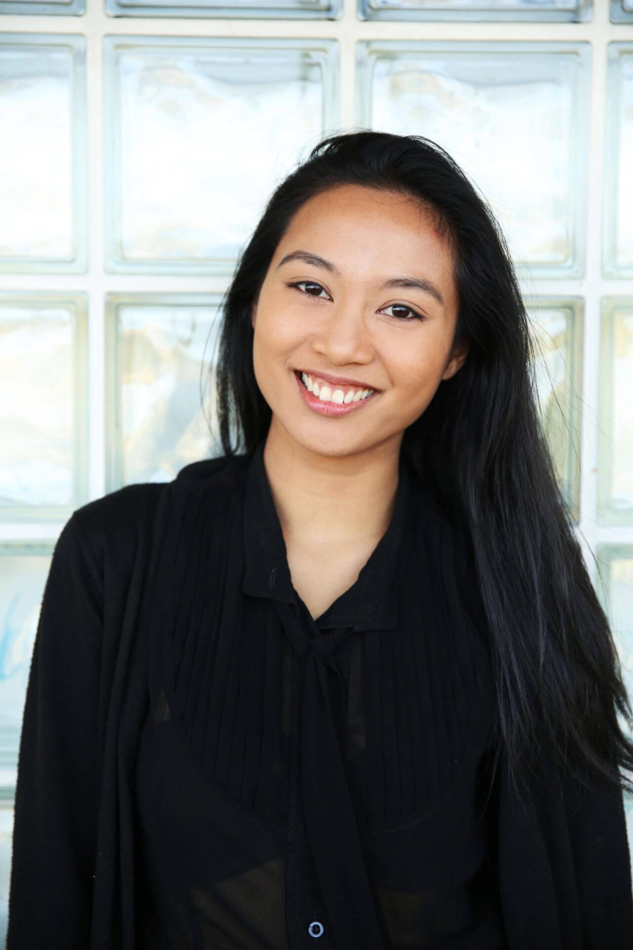 Samantha is smiling at the camera, standing in front of a glass tiled wall. She has long black hair and is wearing a black collared shirt that has a tie at the front and a black cardigan over the top.