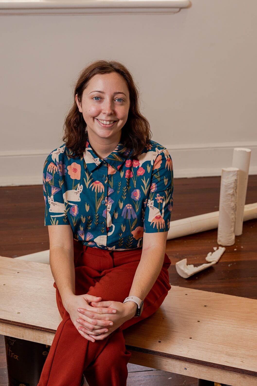 An image of Miranda Johnson sitting on a wooden bench inside a gallery space. Behind her you can see cylindrical plaster structures presented as part of an art installation. She has shoulder-length wavy brown hair and blue eyes and is staring directly at the camera while smiling. She is wearing a blue blouse with a pattern of cats and flowers on it and red pants. Her legs are crossed and she has her hands on her knees.