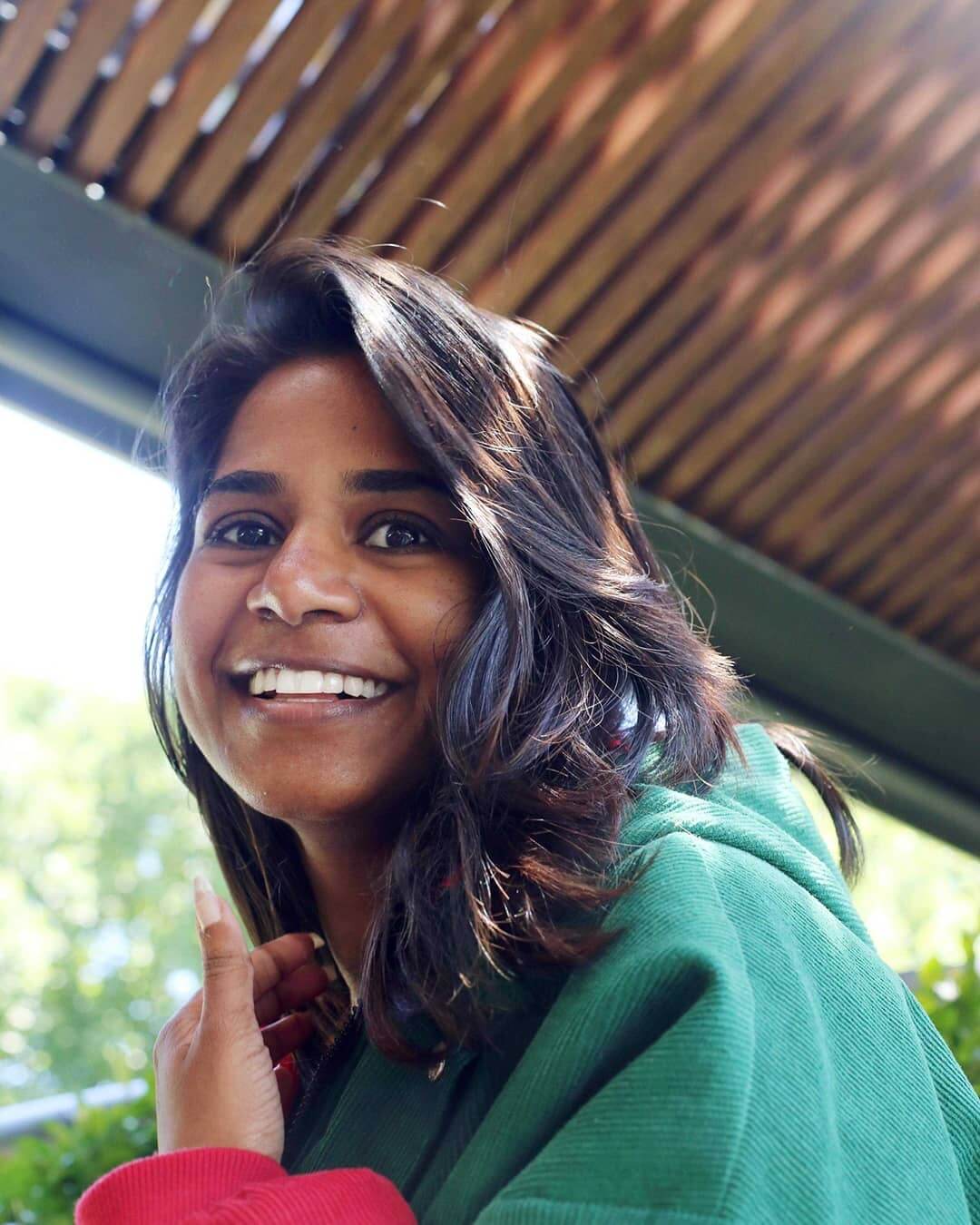 Nithya looks into the camera with happy brown eyes and smiles with her gleaming white teeth. Wearing a teal corduroy jacket with red sleeves, Nithya sits outside under a timber awning with out-of-focus green leafy trees in the background. Nithya brushes her side-parted mid-length dark brown hair to the side with her left hand that has long nails with french tips.