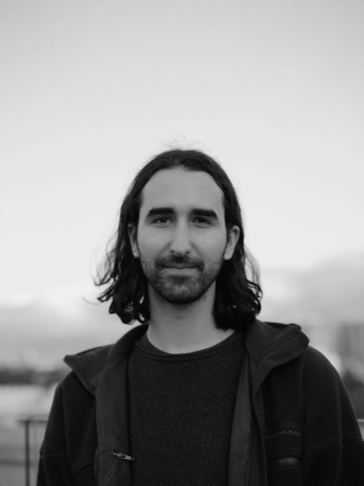 A black and white portrait of Paul Boyé from the chest upwards. The photo was taken outside and the sky is visible behind them. Their hair is dark, wavy and shoulder-length and they have a short black beard and moustache and thick eyebrows. They are looking directly at the camera and slightly smiling. They are wearing a black t-shirt and a dark coloured jacket.