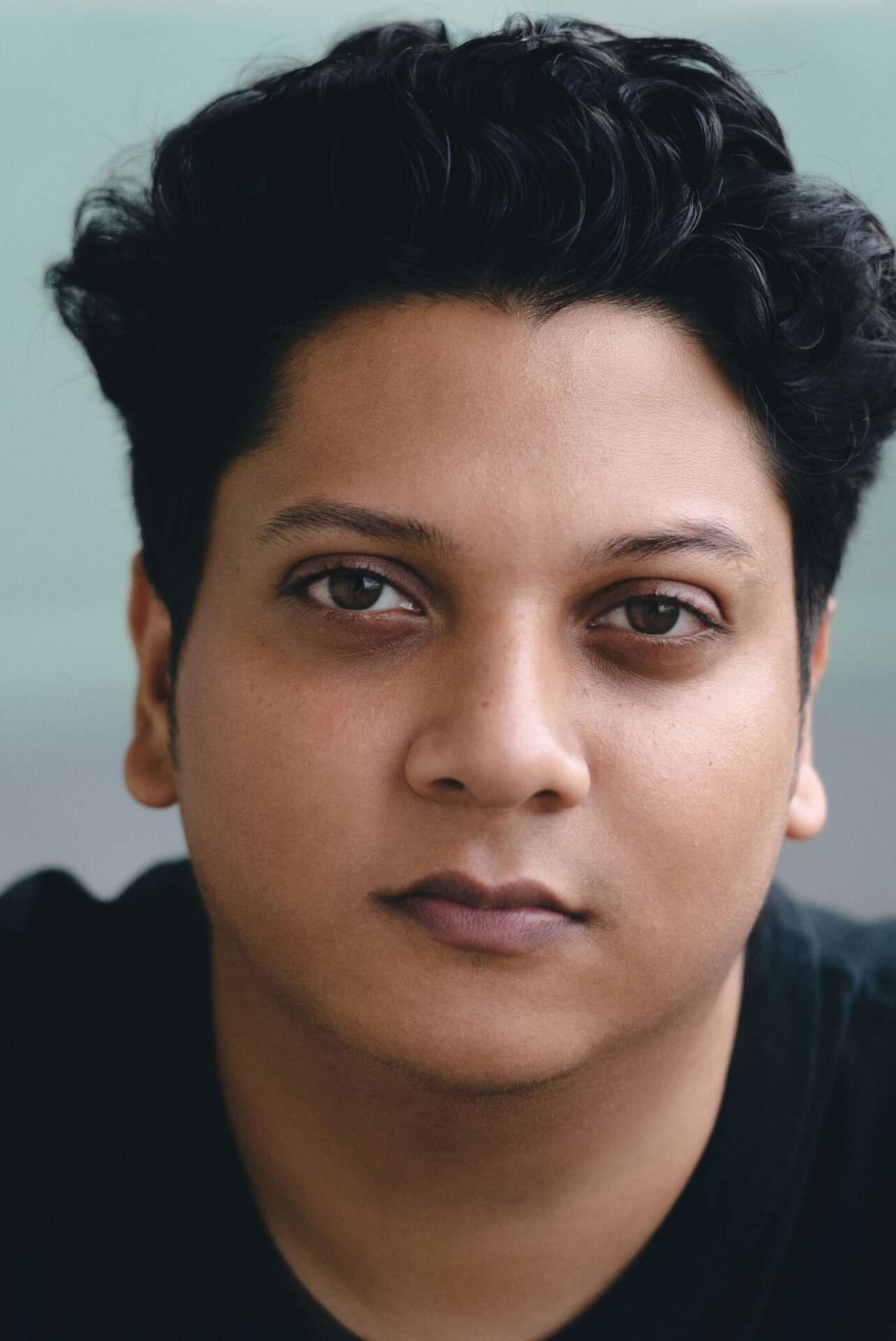 Vishal’s brown eyes look directly into the camera in a tightly cropped portrait with a soft focus. Vishal’s expression appears relaxed and solemn with a slight closed mouth smile. Vishal wears a black shirt and has almost black wavy hair that is long and voluminous on top with shorter sides.